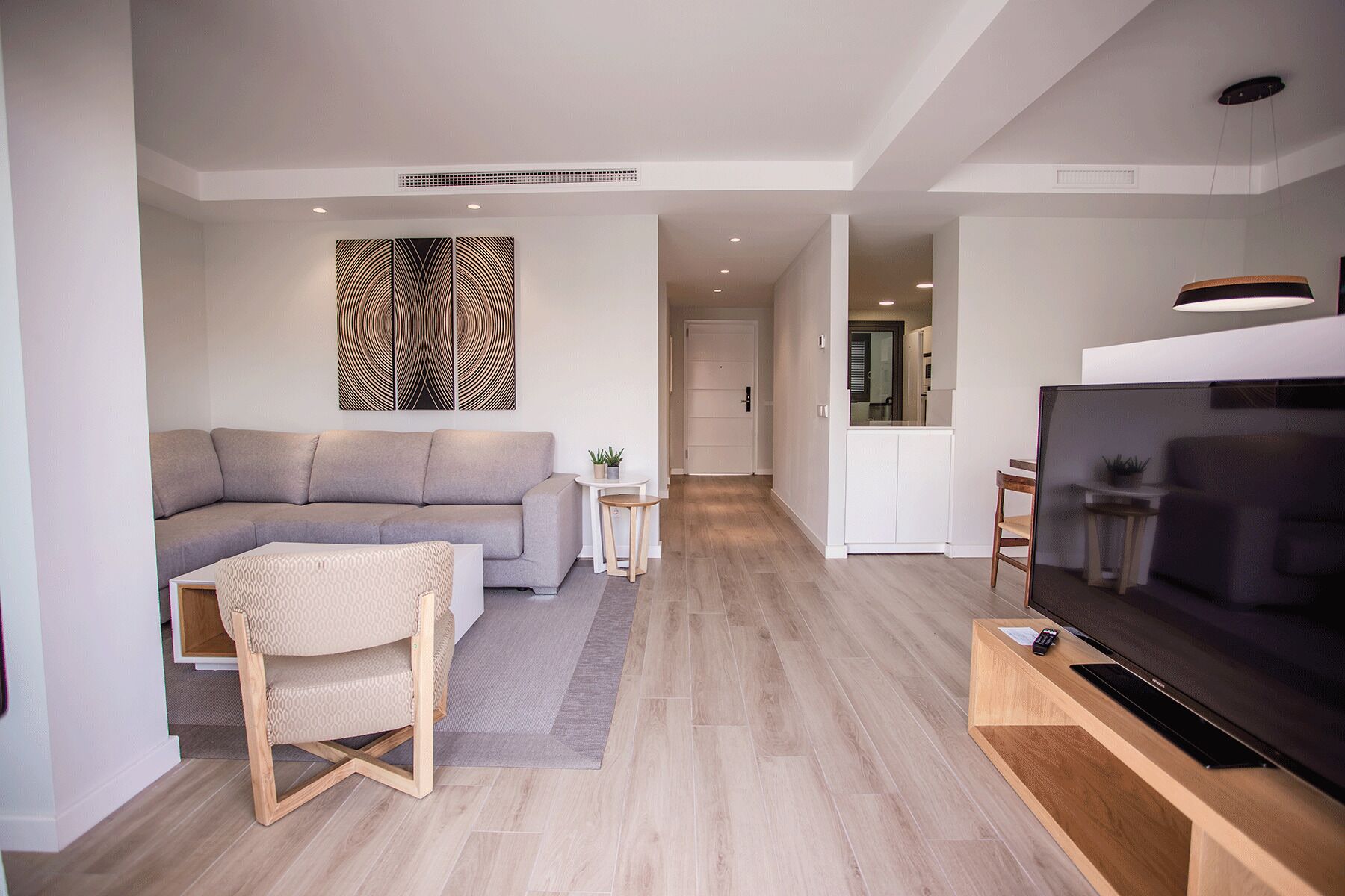 Una sala moderna con un sofá gris, una silla de madera, un televisor de pantalla plana sobre un mueble de madera y una obra de arte colgada en la pared.