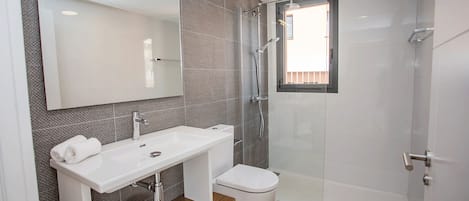 Une salle de bain moderne avec un lavabo blanc, un miroir et des toilettes. Elle comprend un espace douche avec une porte en verre et une petite fenêtre.