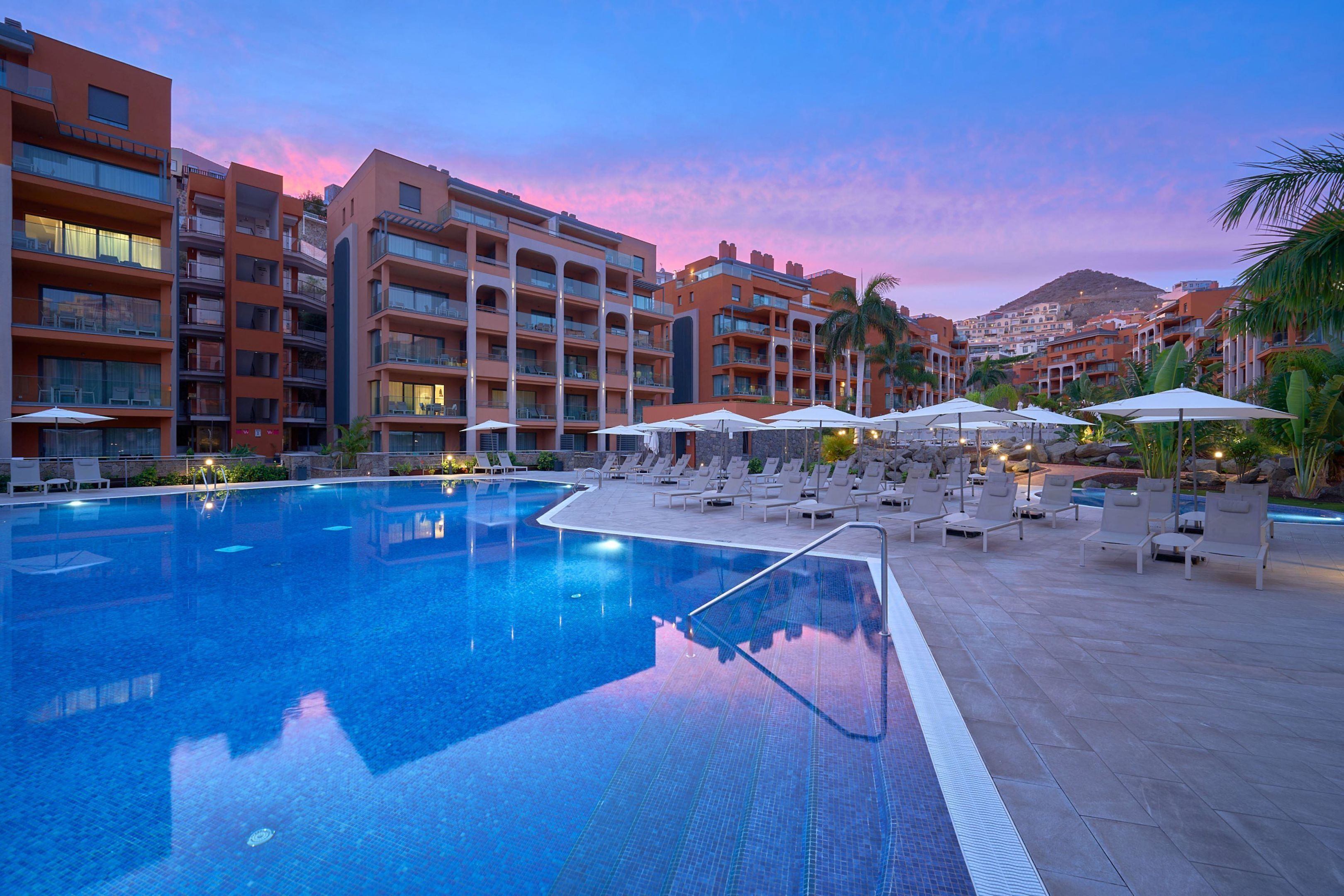 A swimming pool with lounge chairs and umbrellas, surrounded by multi-story buildings with balconies.