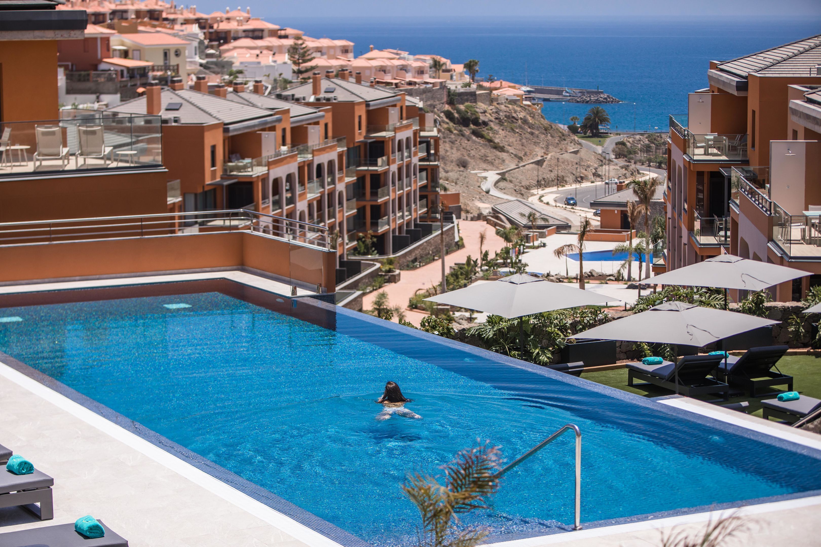 Ein Swimmingpool mit einer Person im Wasser, umgeben von Liegestühlen und Sonnenschirmen, mit Blick auf das Meer und Gebäude im Hintergrund.