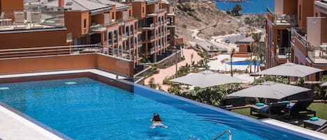 Ein Swimmingpool mit einer Person im Wasser, umgeben von Liegestühlen und Sonnenschirmen, mit Blick auf das Meer und Gebäude im Hintergrund.