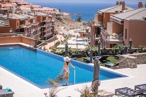 A resort pool area with a clear view of the ocean, a person in a swimsuit, and multiple buildings with balconies. - Arguineguin Park By Servatur VV (Mogan)