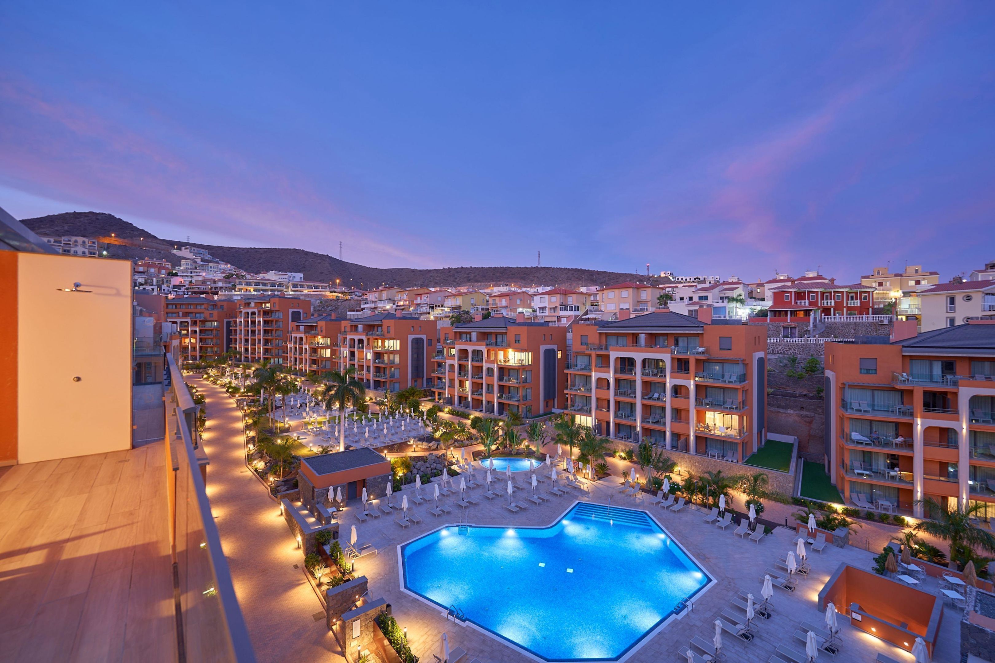 Un resort con una gran piscina, varios balcones y vistas del pueblo circundante.
