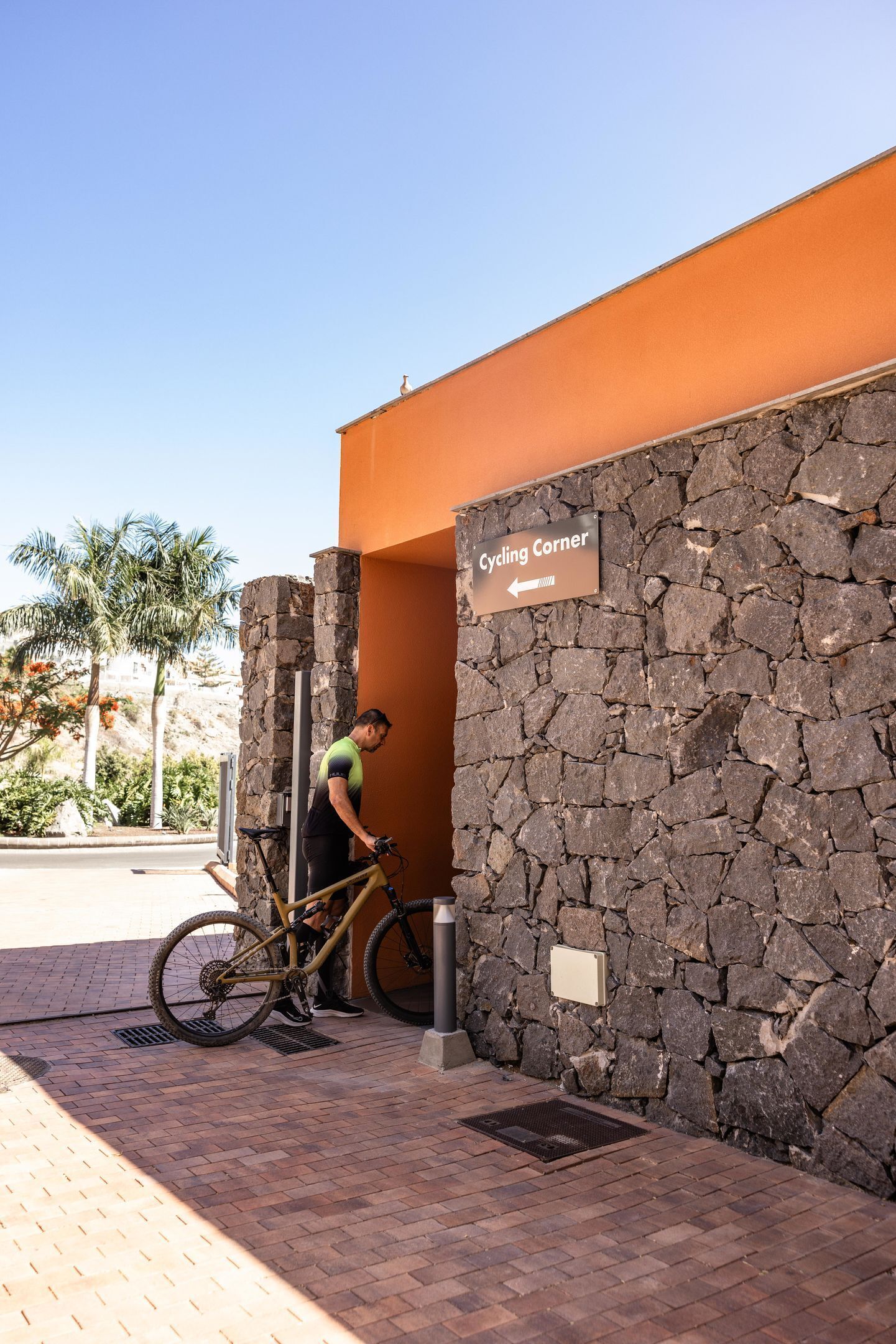 Un muro de piedra con un letrero que dice 'Cycling Corner' y una persona con una bicicleta apoyada en él.