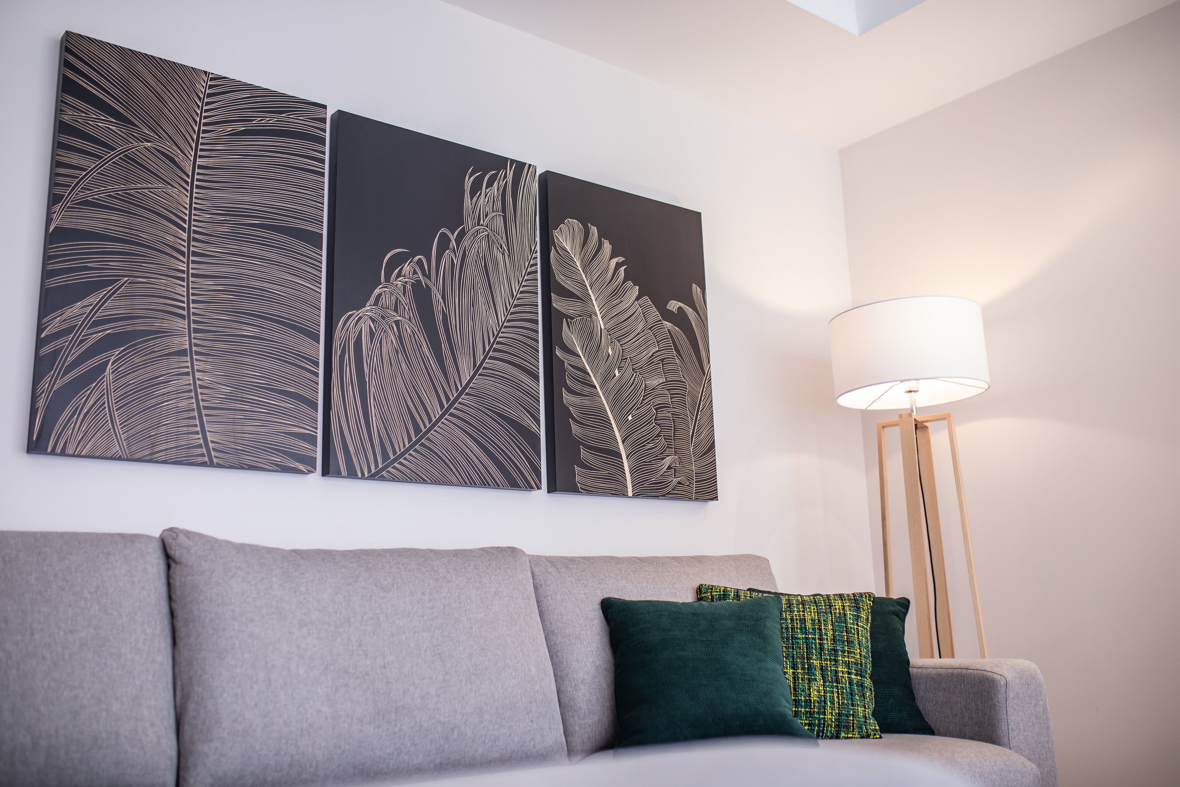 A modern living room with a grey sofa, a floor lamp, and three wall-mounted leaf-patterned artworks.