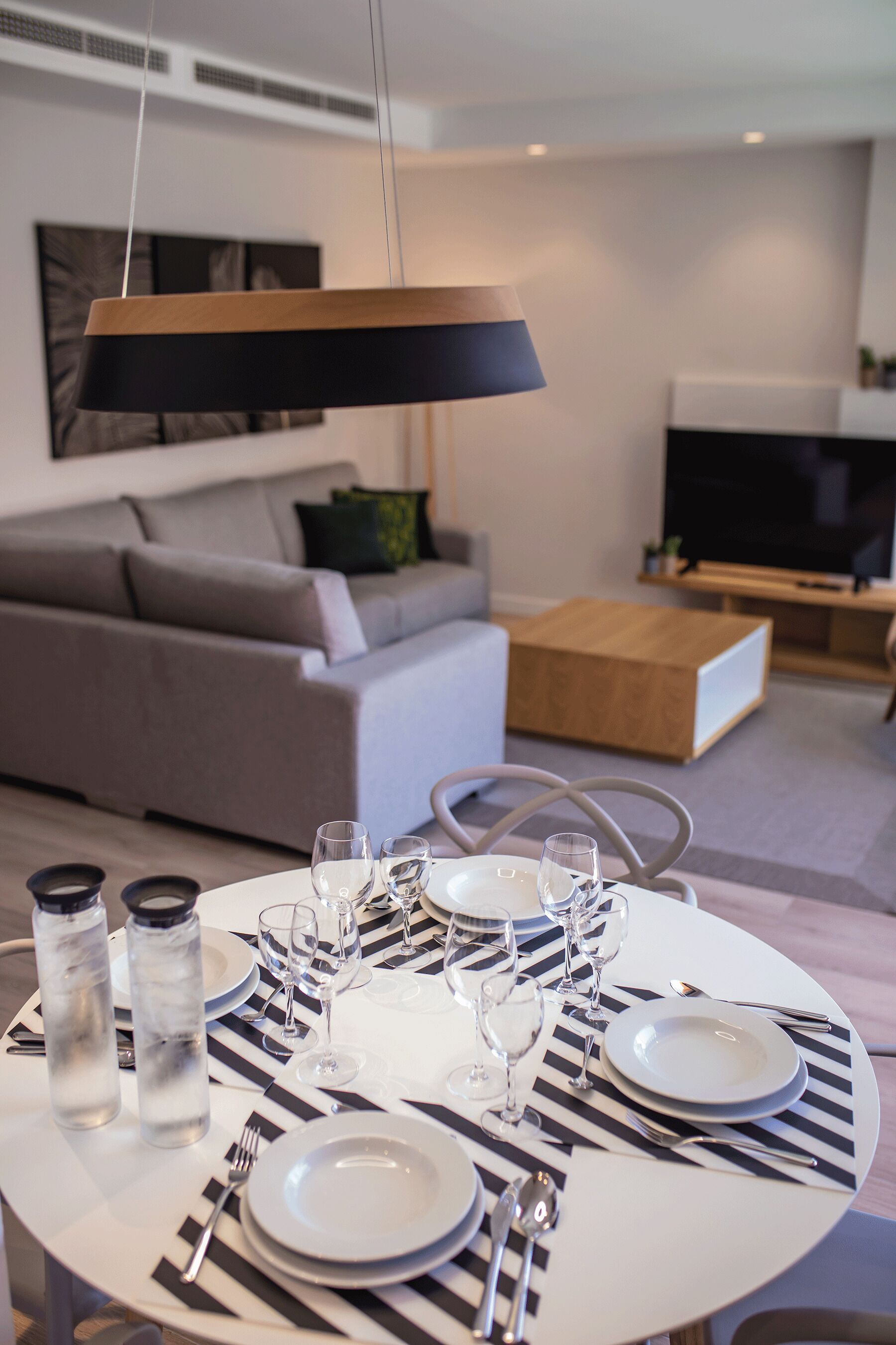 Ein Esszimmer mit Tisch, Tellern, Gläsern, Besteck und Wasserflaschen in einem modernen Wohnzimmer mit Sofa, Couchtisch und Fernseher.