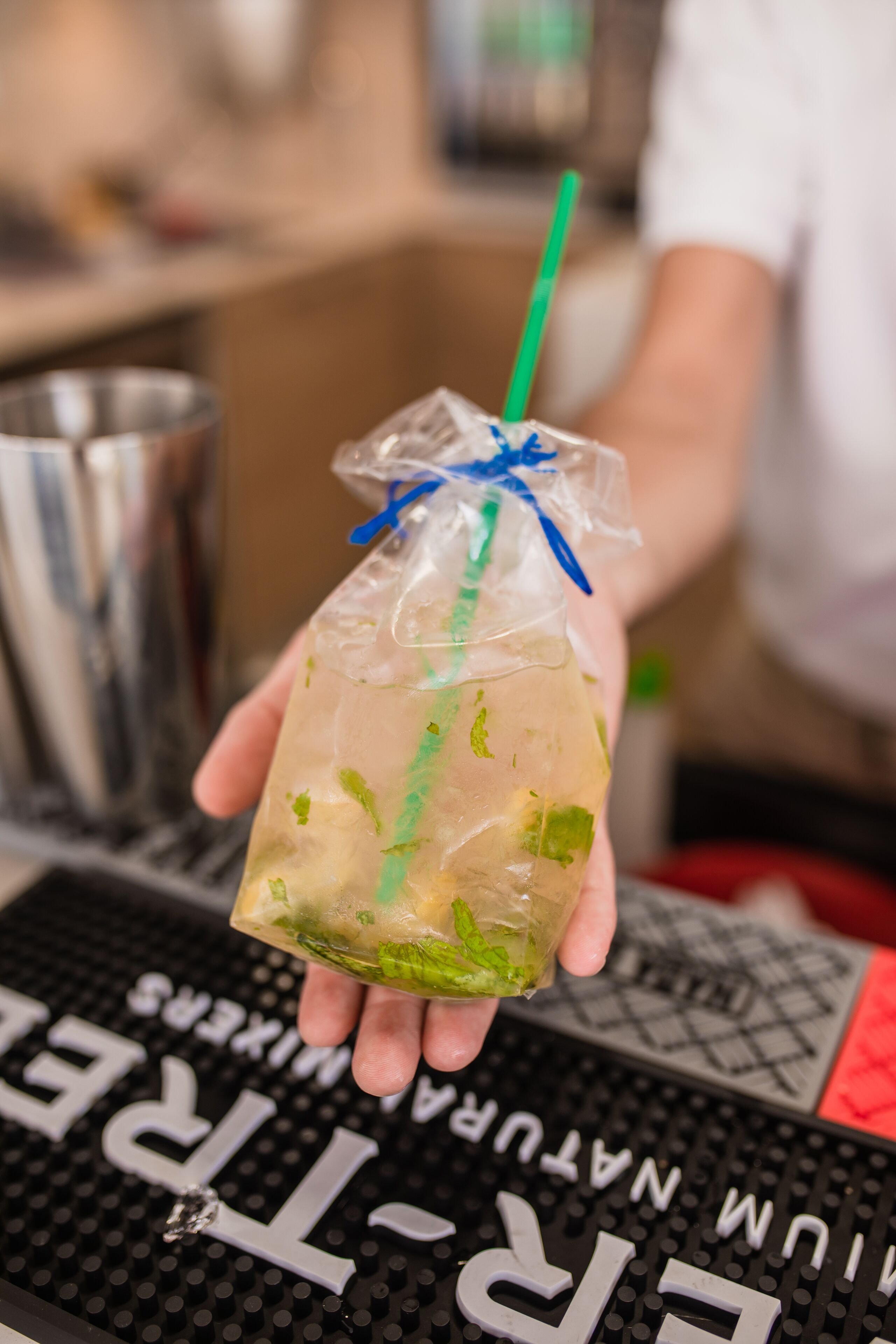 Eine Person hält einen durchsichtigen Plastikbecher mit einem grünen Trinkhalm, gefüllt mit einem Getränk und grüner Dekoration, vor einer Bar mit einem Metall-Cocktailshaker und einer Tastatur mit Beschriftung.