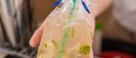 Eine Person hält einen durchsichtigen Plastikbecher mit einem grünen Trinkhalm, gefüllt mit einem Getränk und grüner Dekoration, vor einer Bar mit einem Metall-Cocktailshaker und einer Tastatur mit Beschriftung.