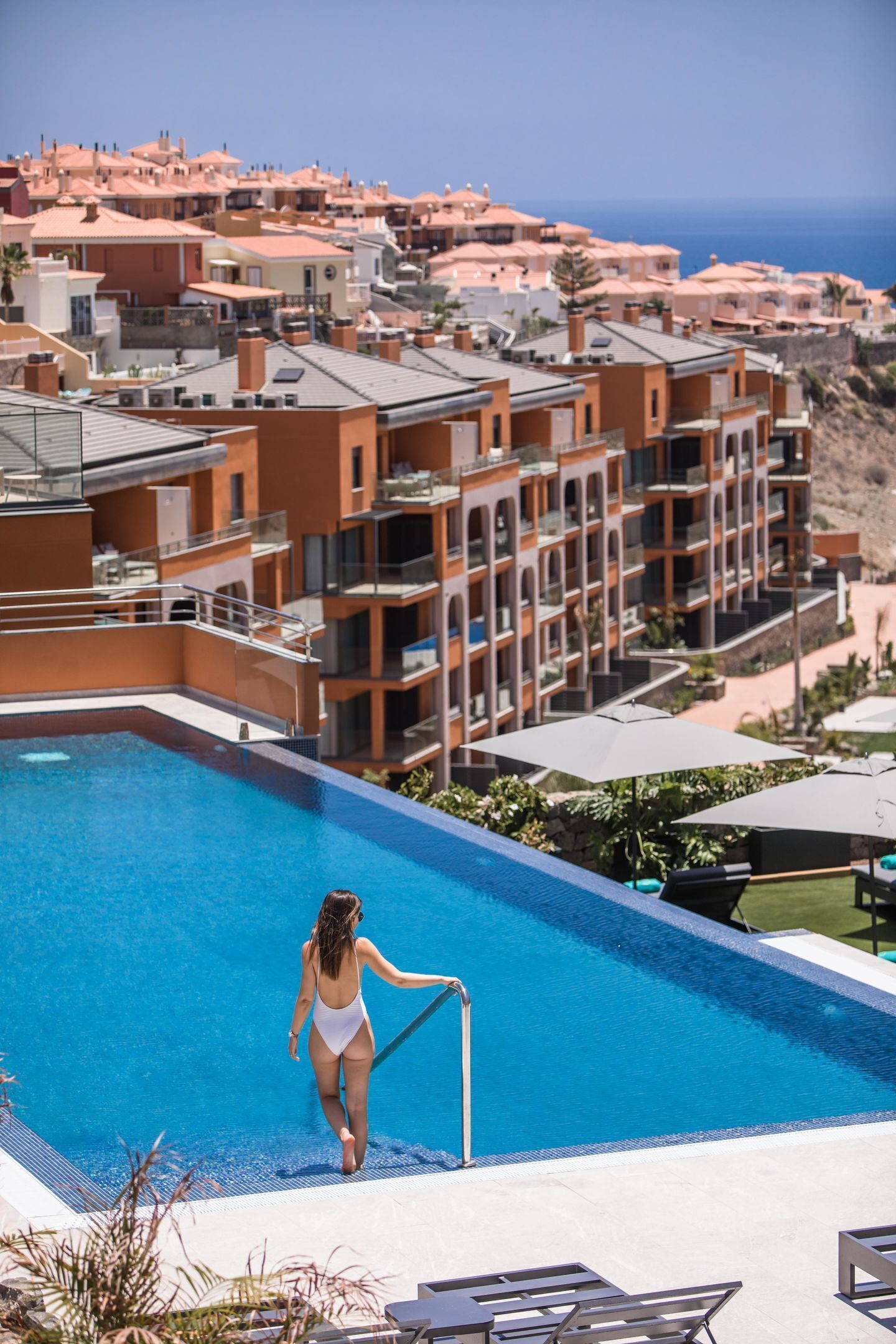 A resort with a swimming pool, a woman in a swimsuit, and a view of the ocean and buildings in the background.