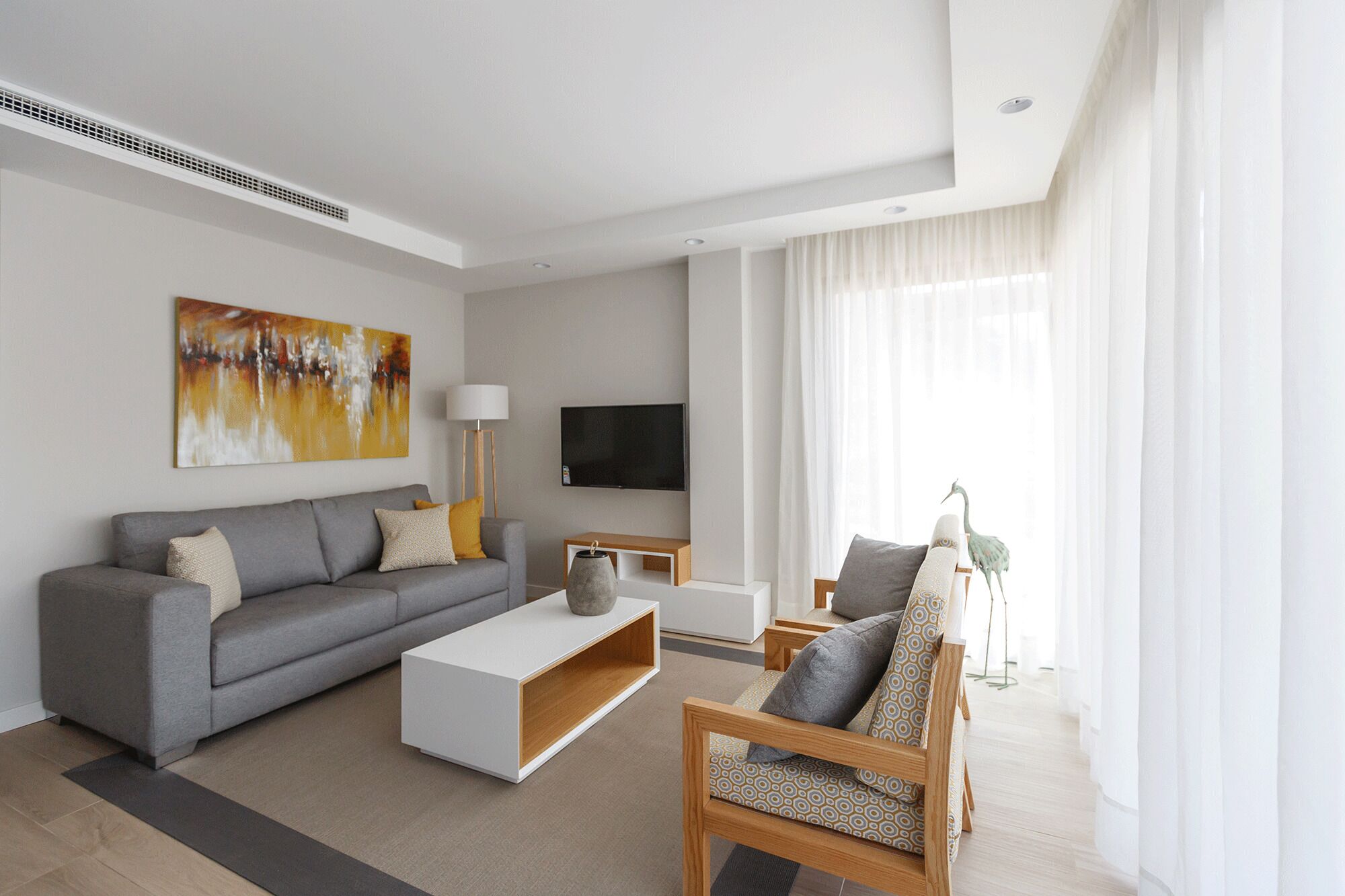 A modern living room with a grey sofa, white coffee table, and a wooden chair. A flat-screen TV is mounted on the wall, and there's a floor lamp beside the sofa.
