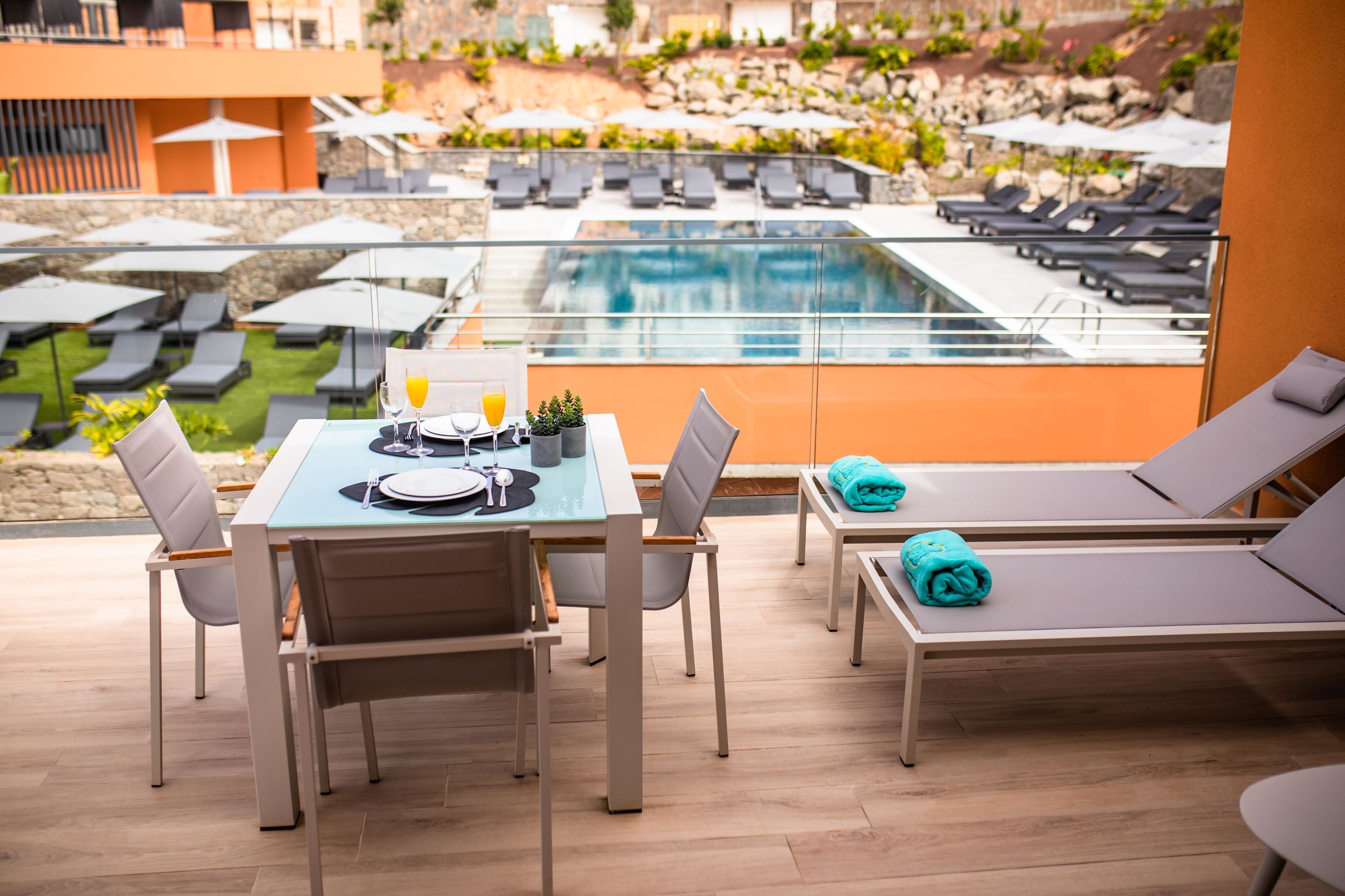 Una terraza con mesa y sillas para dos personas, con vistas a una zona de piscina con sillones y sombrillas.
