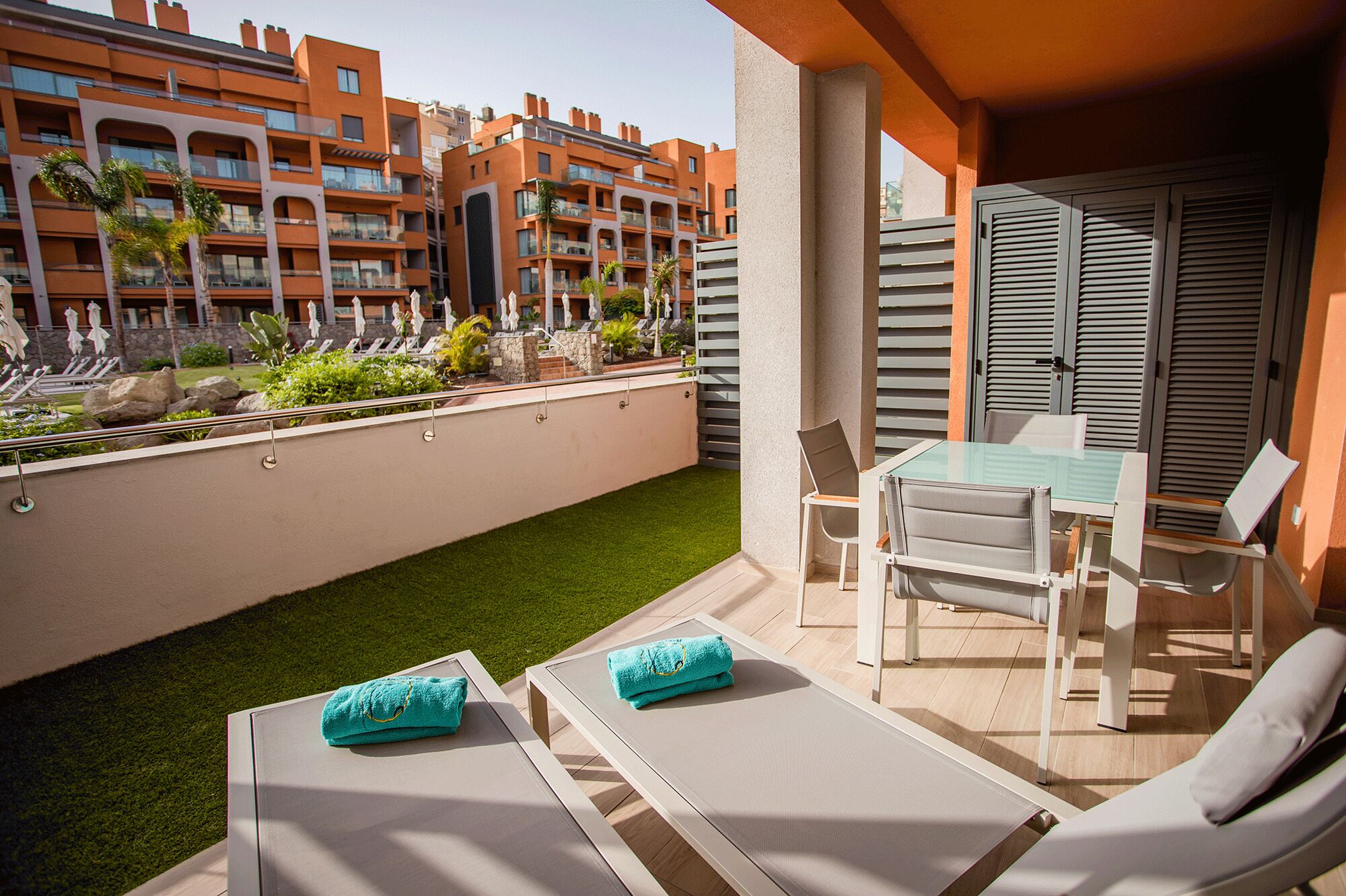 Un balcó amb taula i caires de vidre, una petita zona de jardí amb gespa artificial, i vista a edificis veïnals.