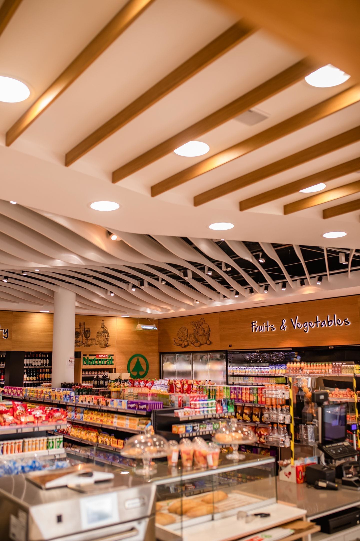 Interior de un supermercado con una sección de 'Fruits & Vegetables', vigas de madera en el techo y una variedad de productos envasados en estantes.