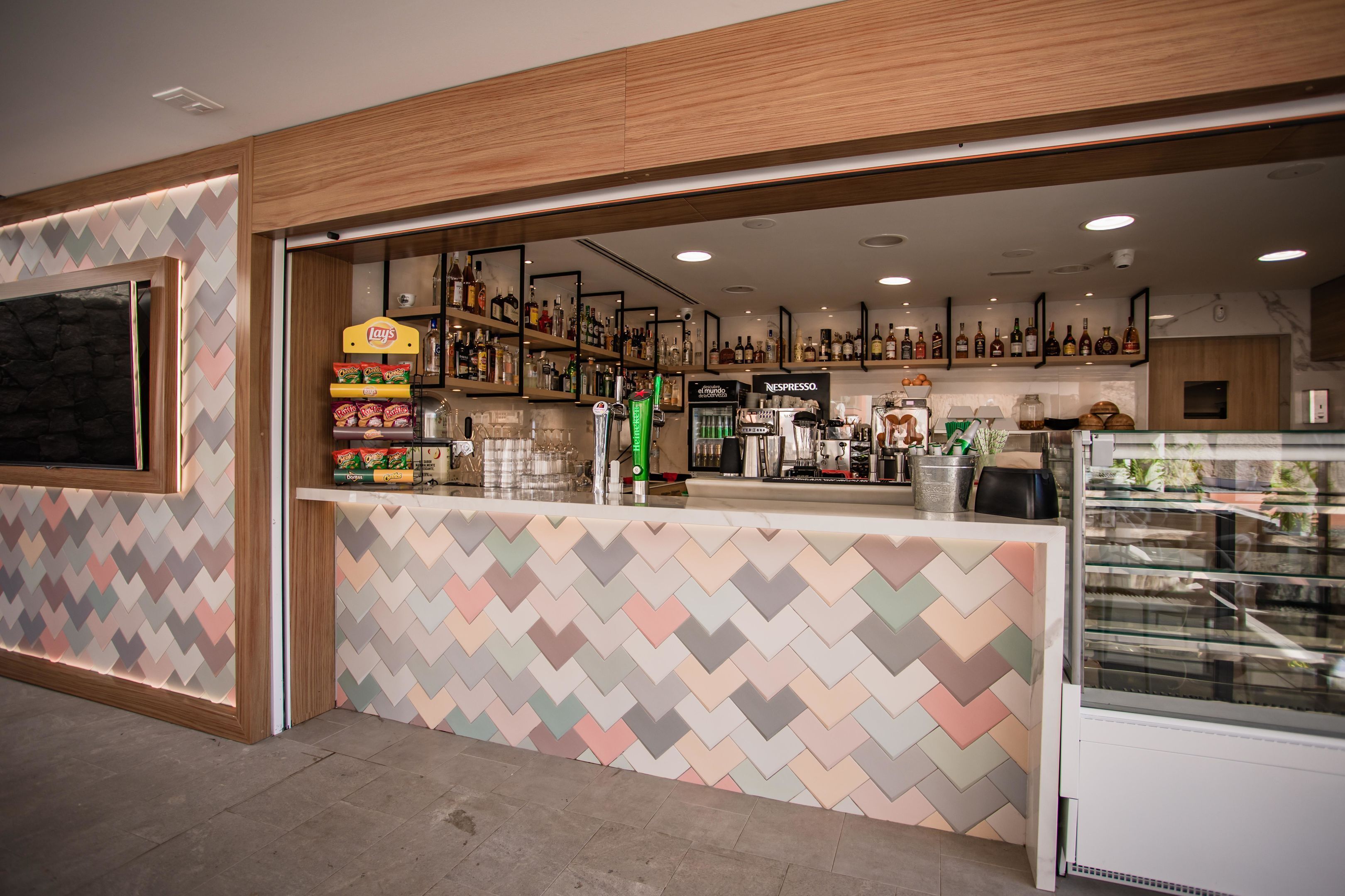 A modern bar area with a herringbone patterned wall, shelves stocked with snacks and beverages, and a display case with pastries.