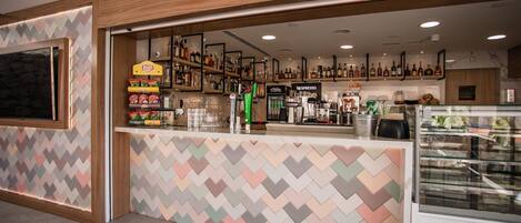 A modern bar area with a herringbone patterned wall, shelves stocked with snacks and beverages, and a display case with pastries.
