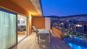 A balcony with a dining table and chairs, a glass door leading to a room with a bed, and a view of a swimming pool and buildings in the background.