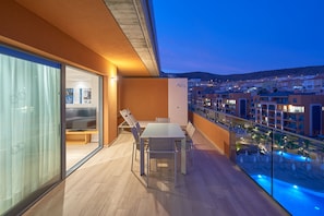 Un balcón con mesa y sillas, un ventanal de vidrio que da a una habitación con cama, y vistas a una piscina y edificios al fondo.