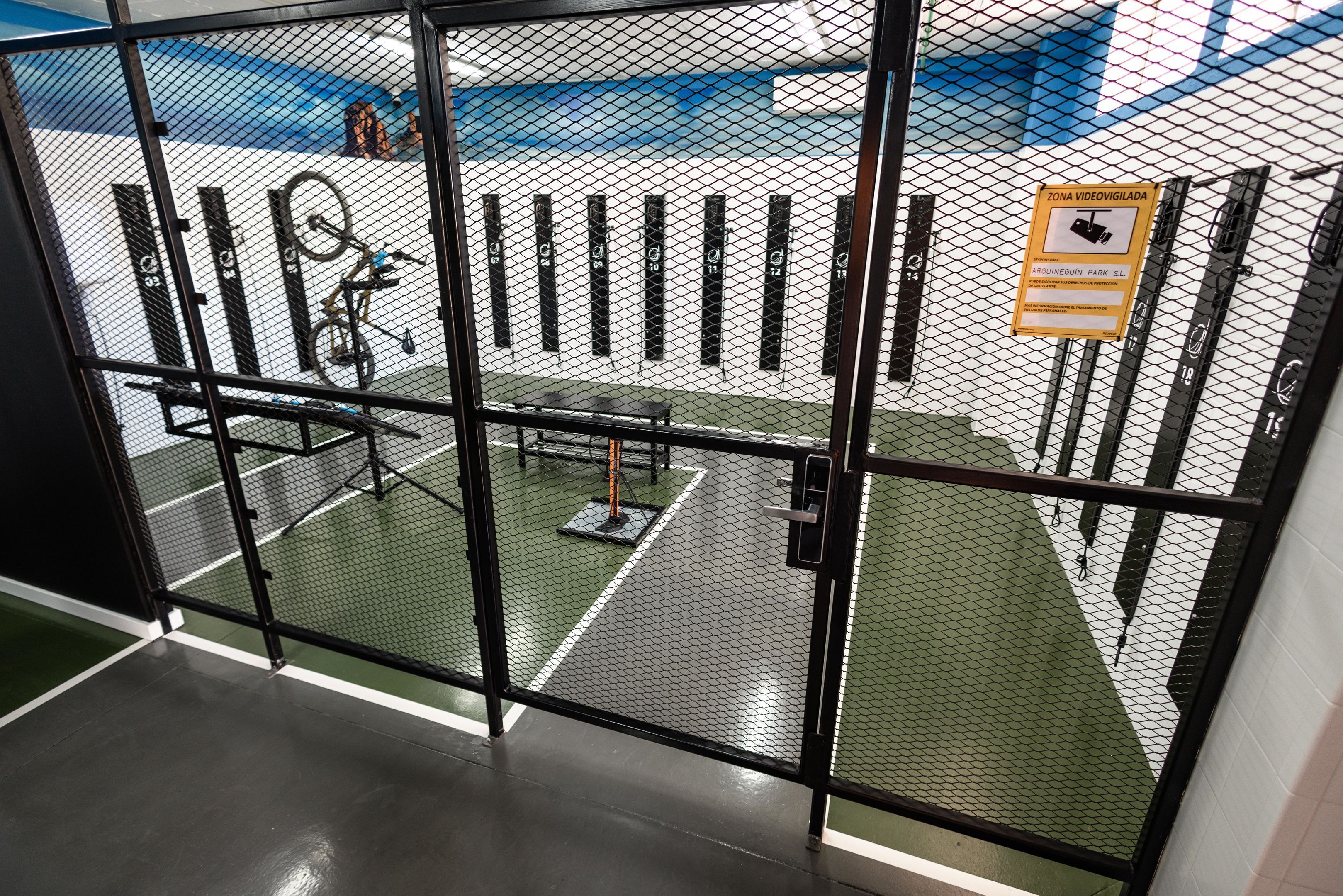 A view through a metal gate into a gym area with exercise equipment and a sign indicating surveillance.