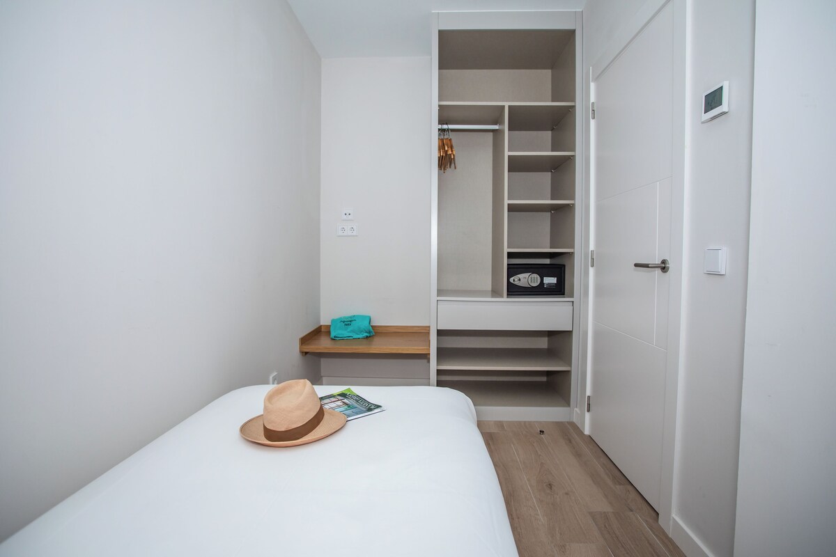Une chambre à coucher avec un lit, une table de chevet en bois, un chapeau, un magazine, une étagère avec une radio et une armoire contenant des vêtements suspendus.