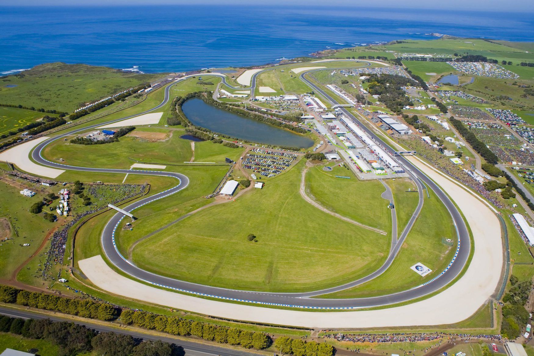 An aerial view of a racetrack with multiple curves, spectator areas, and a large body of water nearby.