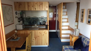 A compact kitchen with wooden cabinets, a dining table with chairs, a small refrigerator, and a microwave. There is a staircase leading to a bedroom with a bed and a blue plaid sofa. - Duplex Apartment in Arc 1800, Aiguille Grive 3, 40m2, ski in/out (Bourg-Saint-Maurice)