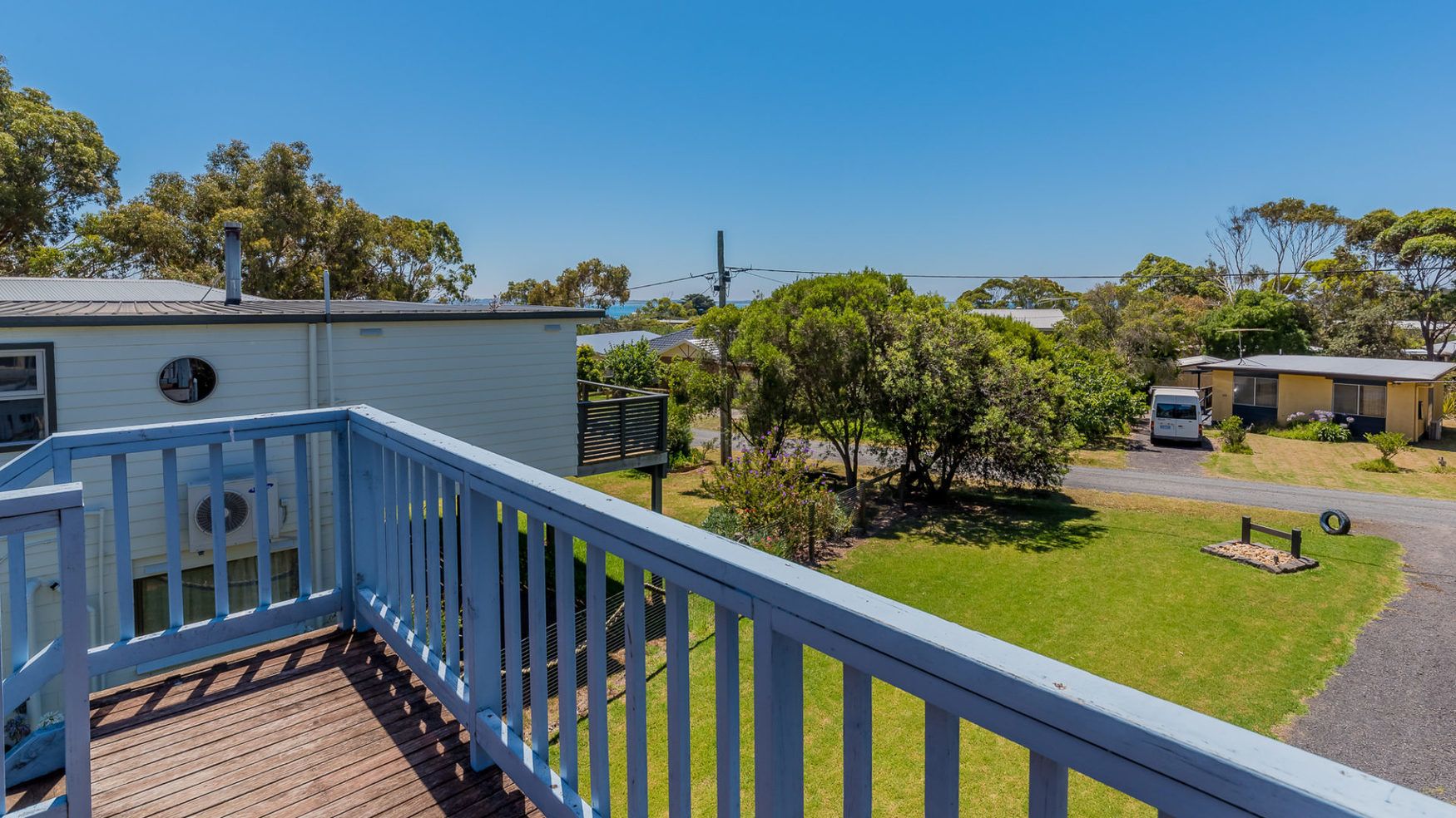 A wooden deck with a blue railing overlooking a grassy area with trees and a building in the background.
