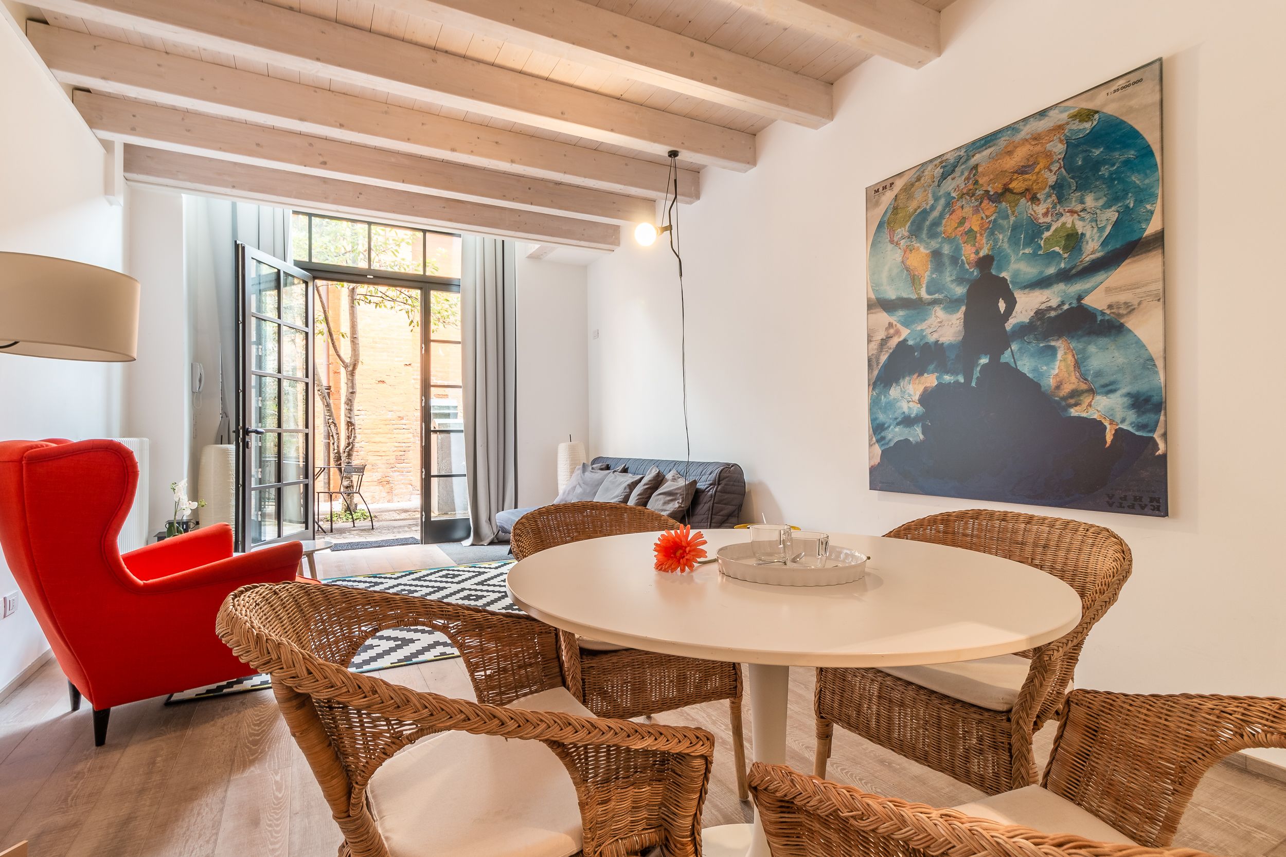 A modern living room with a round dining table, wicker chairs, a red armchair, and a large abstract painting on the wall.