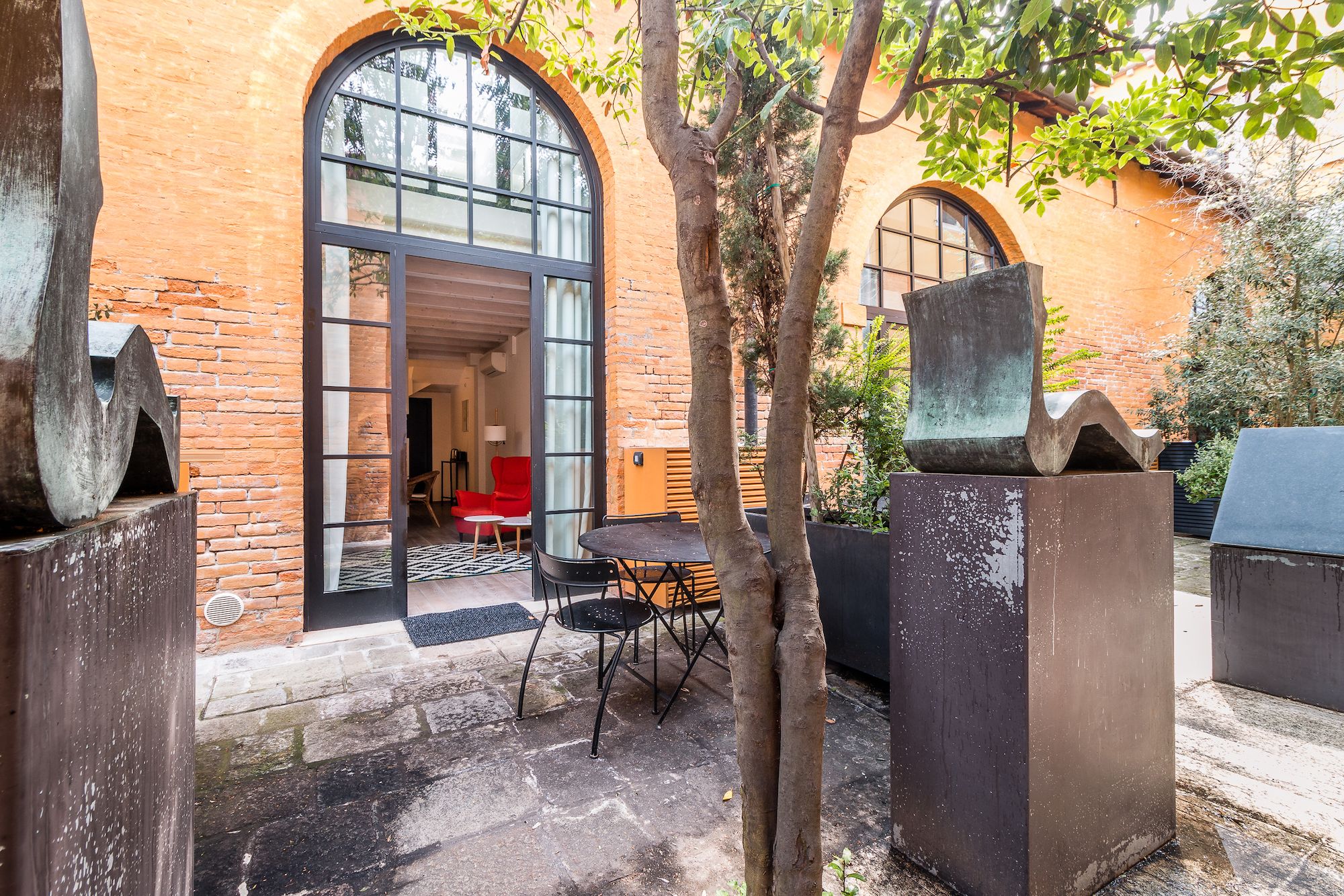 Un espace extérieur aménagé avec une table et des chaises, dont une chaise rouge, et une grande fenêtre donnant sur l’intérieur du bâtiment.