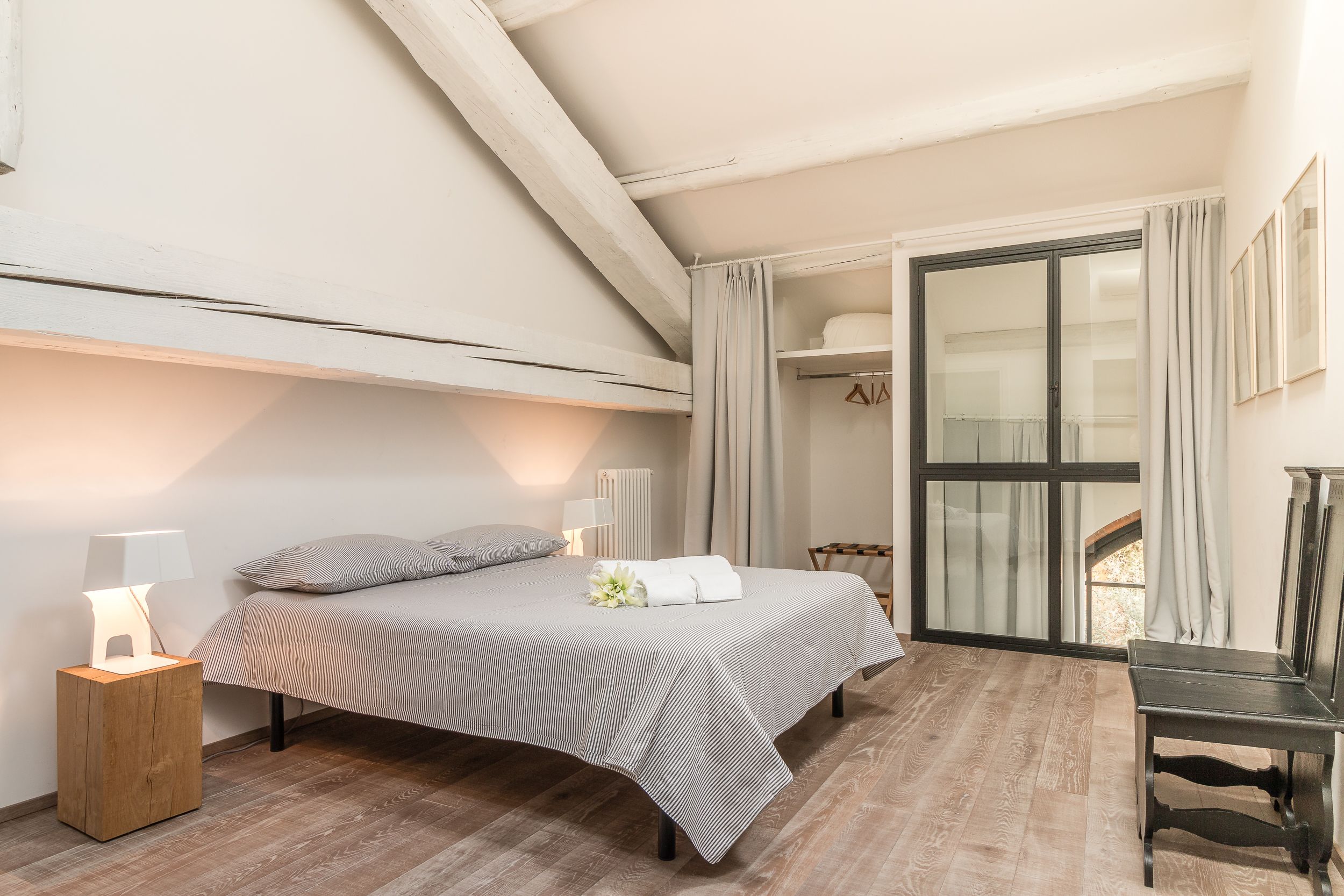 Un dormitorio con cama, lámparas de noche, una mesita de noche de madera, un banco y vistas a través de un ventanal.