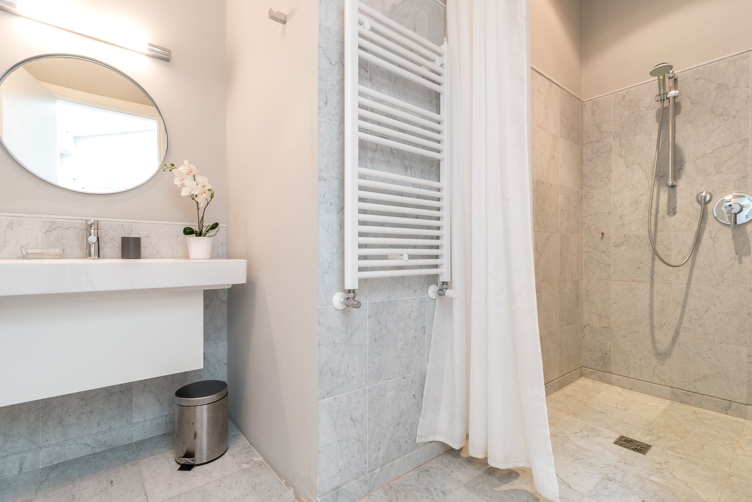 A modern bathroom with a white vanity, a round mirror, a white towel rack, a shower with a handheld showerhead, and a trash can.