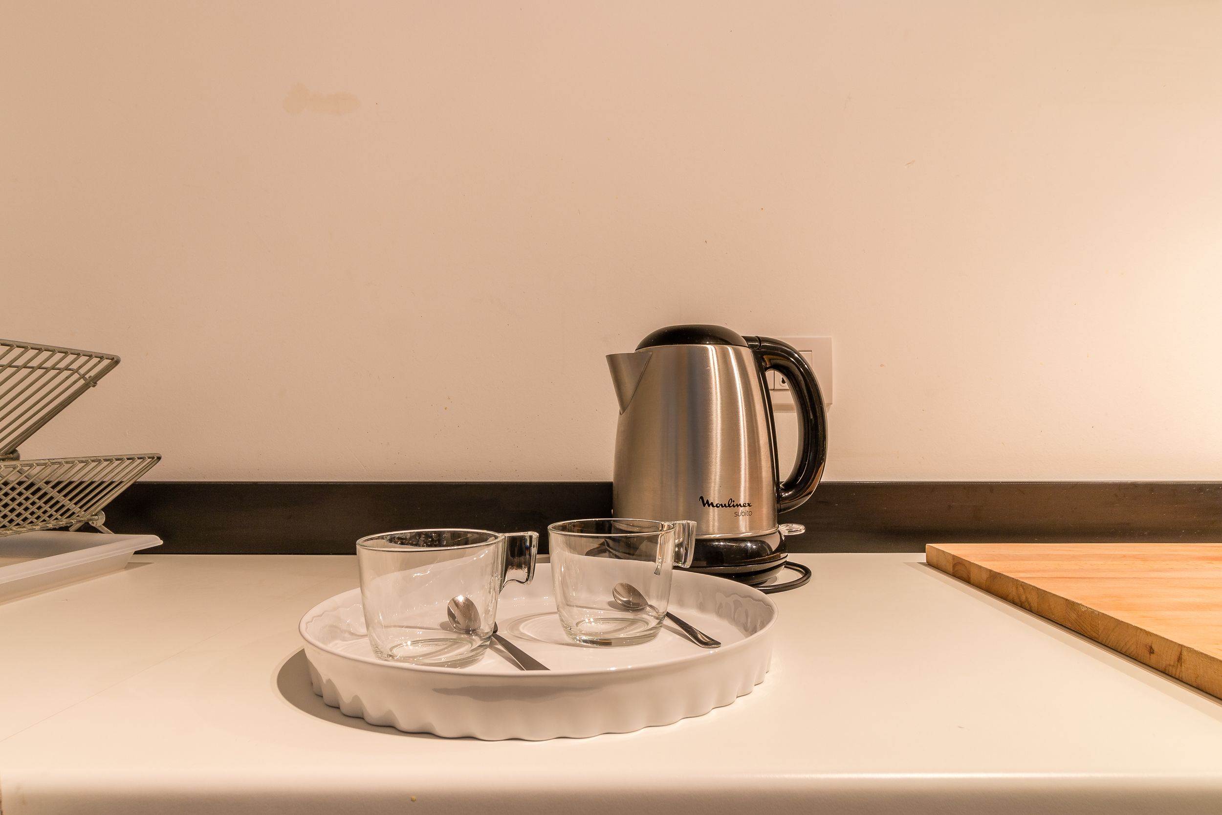 A kitchen counter with a coffee maker, a tray with two glasses and spoons, and a wooden cutting board.
