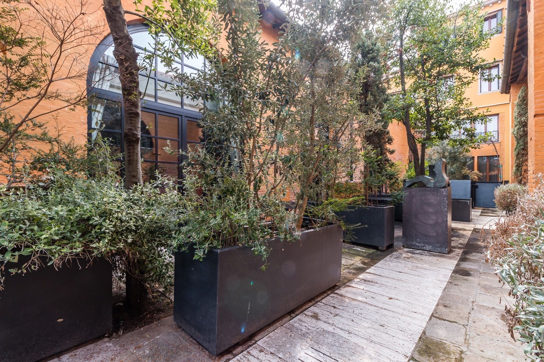 A courtyard with potted plants, a wooden walkway, and a building with large windows and a curved glass facade.