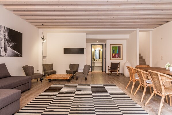 Uma sala de estar moderna com um sofá cinza, duas poltronas, uma mesa de centro e uma mesa de jantar com cadeiras de vime. Um tapete listrado em preto e branco está no chão.