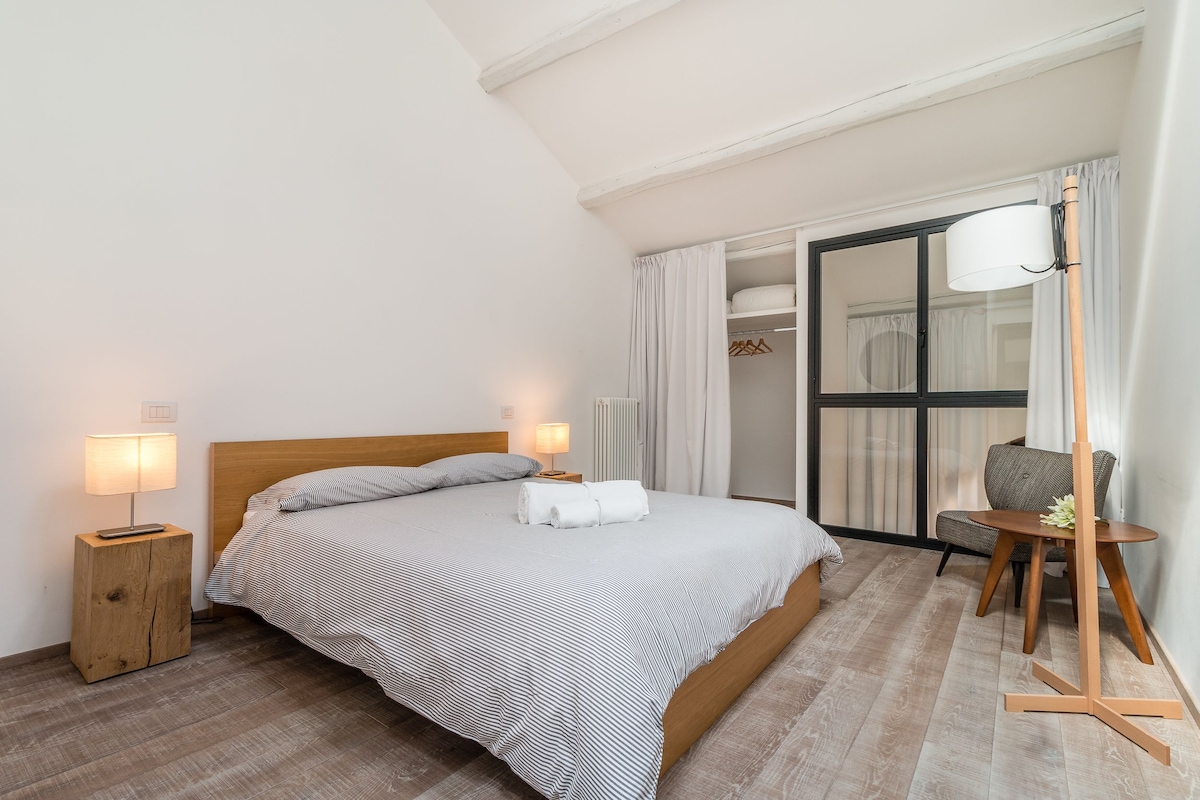 Un lit bien fait, recouvert d’une couvre-lit à rayures, deux lampes de chevet, une table de chevet en bois, une chaise, une table avec une lampe, et une vue sur un placard à portes coulissantes en verre.