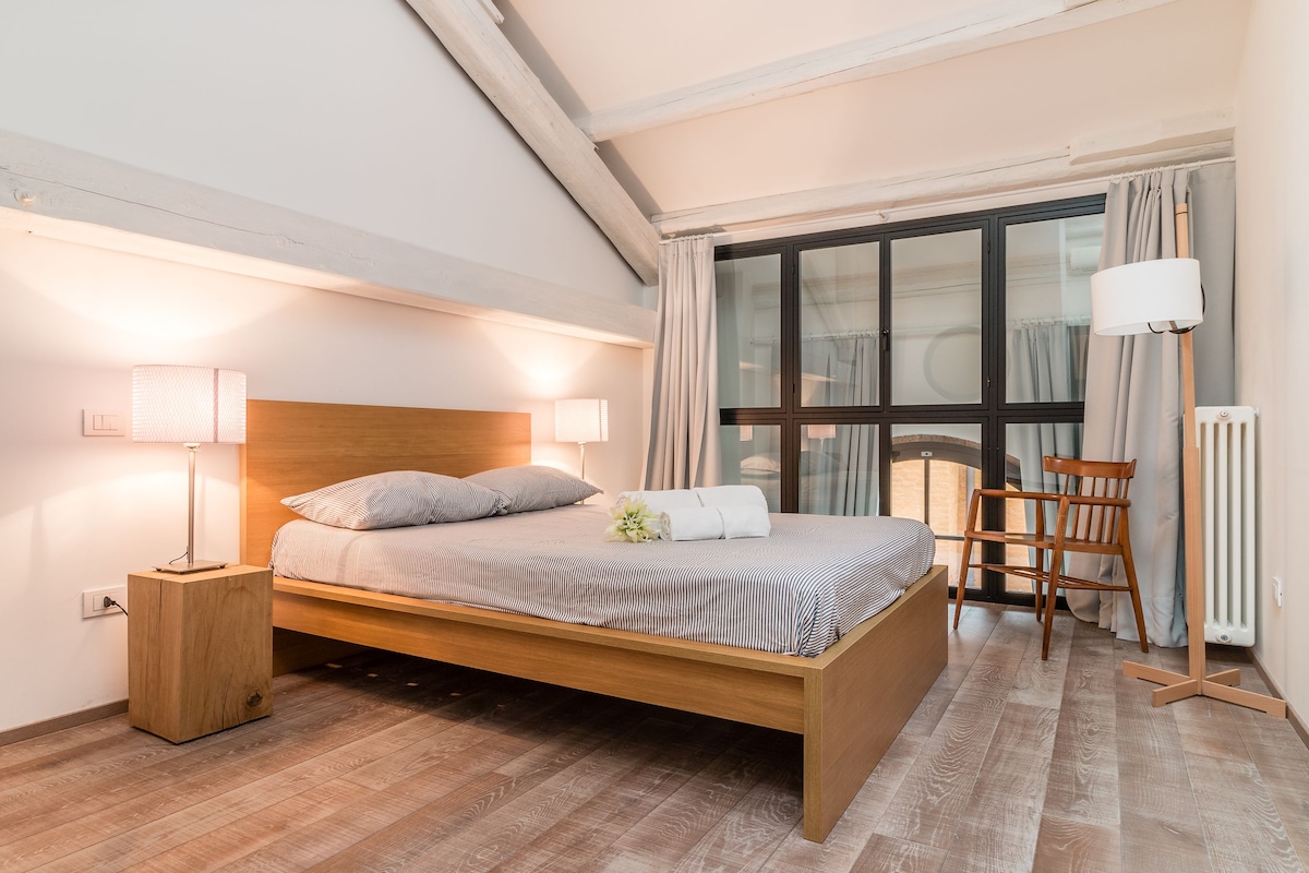 Un lit bien fait, avec une tête de lit en bois, une table de chevet avec une lampe, une chaise et une fenêtre avec des rideaux.