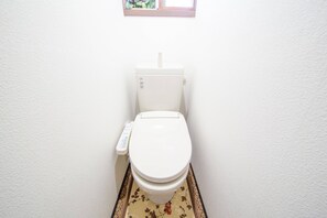 A white toilet with a remote control, a window above it, and a floral-patterned mat. - NexStay HOUSE Osaka West (Osaka)