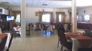A dining area with multiple tables and chairs, a television on the wall, and a kitchen area in the background.