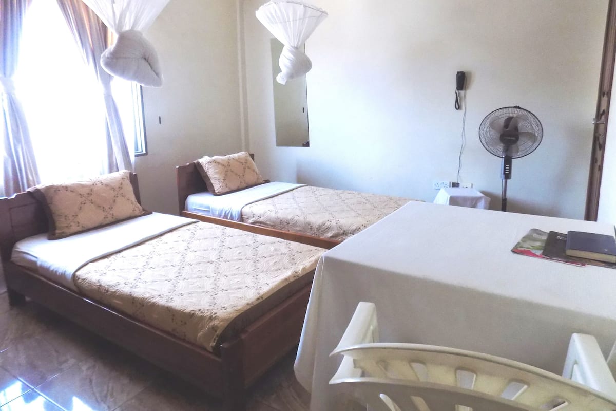 A room with two single beds, a table, a chair, a standing fan, and a window with curtains.