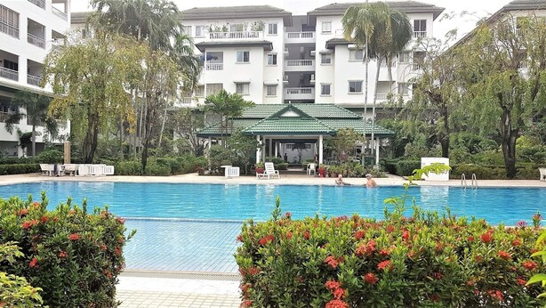 A swimming pool surrounded by lush greenery and a multi-story building with balconies. - Luxury 1 bed Corner Condo (Pattaya)