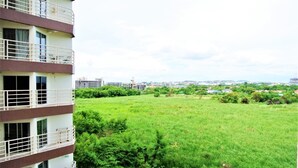 A multi-story building with balconies, a grassy field, and distant buildings. - Lovely Jomtien Beach Apartment (Pattaya)