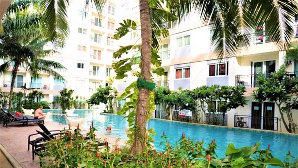 A swimming pool surrounded by lush greenery and palm trees, with a building in the background. - Park Lane Resort Jomtien Beach (Pattaya)