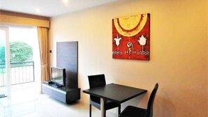 Een moderne woonkamer met een flatscreen televisie, een eettafel met twee stoelen, een groot abstract schilderij aan de muur en een balkon met uitzicht op groen.