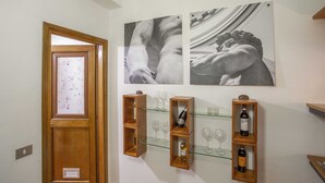 A hallway with wooden shelves displaying wine bottles and glasses, a wooden door with a frosted glass panel, and two black-and-white photographs on the wall.