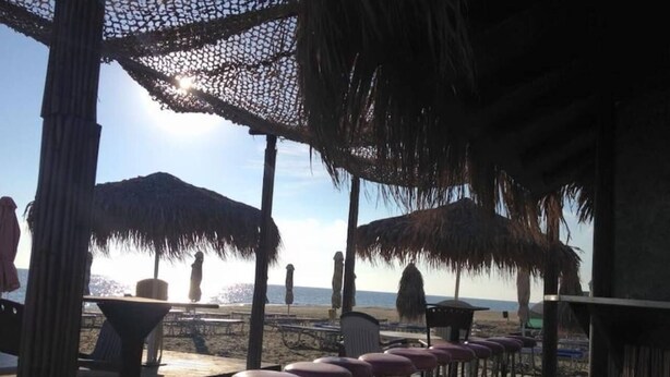 An outdoor seating area with thatched umbrellas, wooden tables, and bar stools, overlooking a beach. - Luxury oasis with garden near beach and airport (Tersefanou)