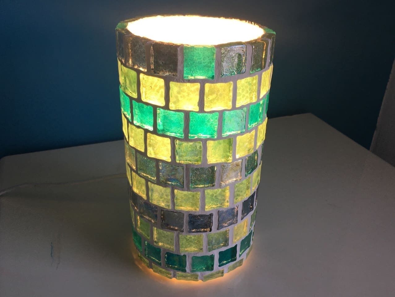 Un objet cylindrique orné d’un motif en mosaïque, vraisemblablement une lampe ou un objet décoratif, placé sur une surface plane devant un fond sombre.