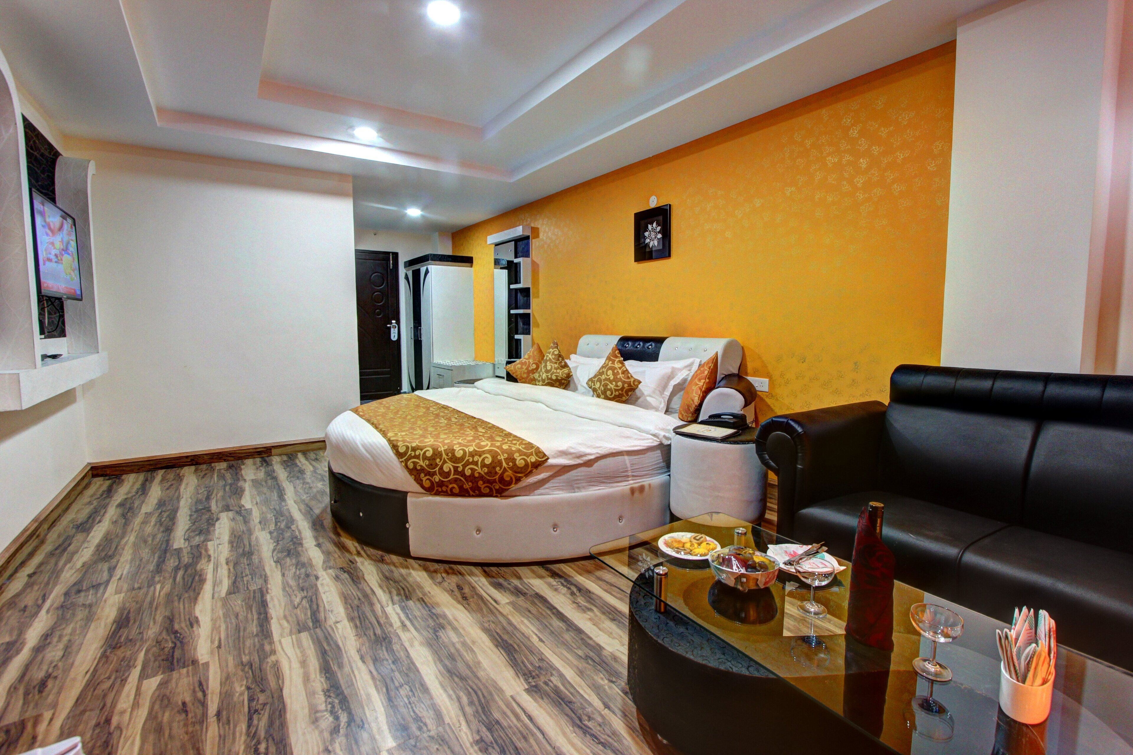 A hotel room with a bed, a black leather sofa, a glass coffee table with items on it, and a television mounted on the wall.