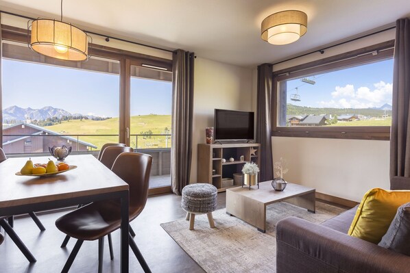 A modern living room with a dining area, large windows with mountain views, a flat-screen TV, and a cozy seating arrangement. - Chalet in French Alps with Premium Comfort (Hauteluce)