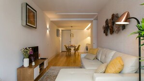 A modern living room with a white sofa, a wooden side table, and a TV. There is a dining area with a table and chairs in the background. - JOIVY Sunny 1-bed flat w/terrace&sea view in Baixa,3mins to Arco da Rua Augusta (Lisboa)