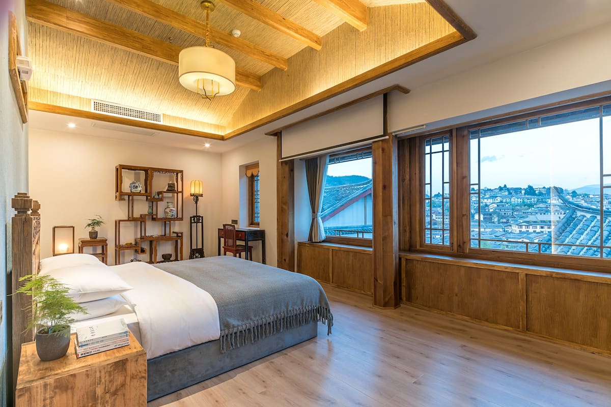 Une chambre spacieuse avec un grand lit, des poutres apparentes en bois au plafond, une vue sur le paysage urbain à travers de grandes fenêtres et un petit bureau avec une chaise.