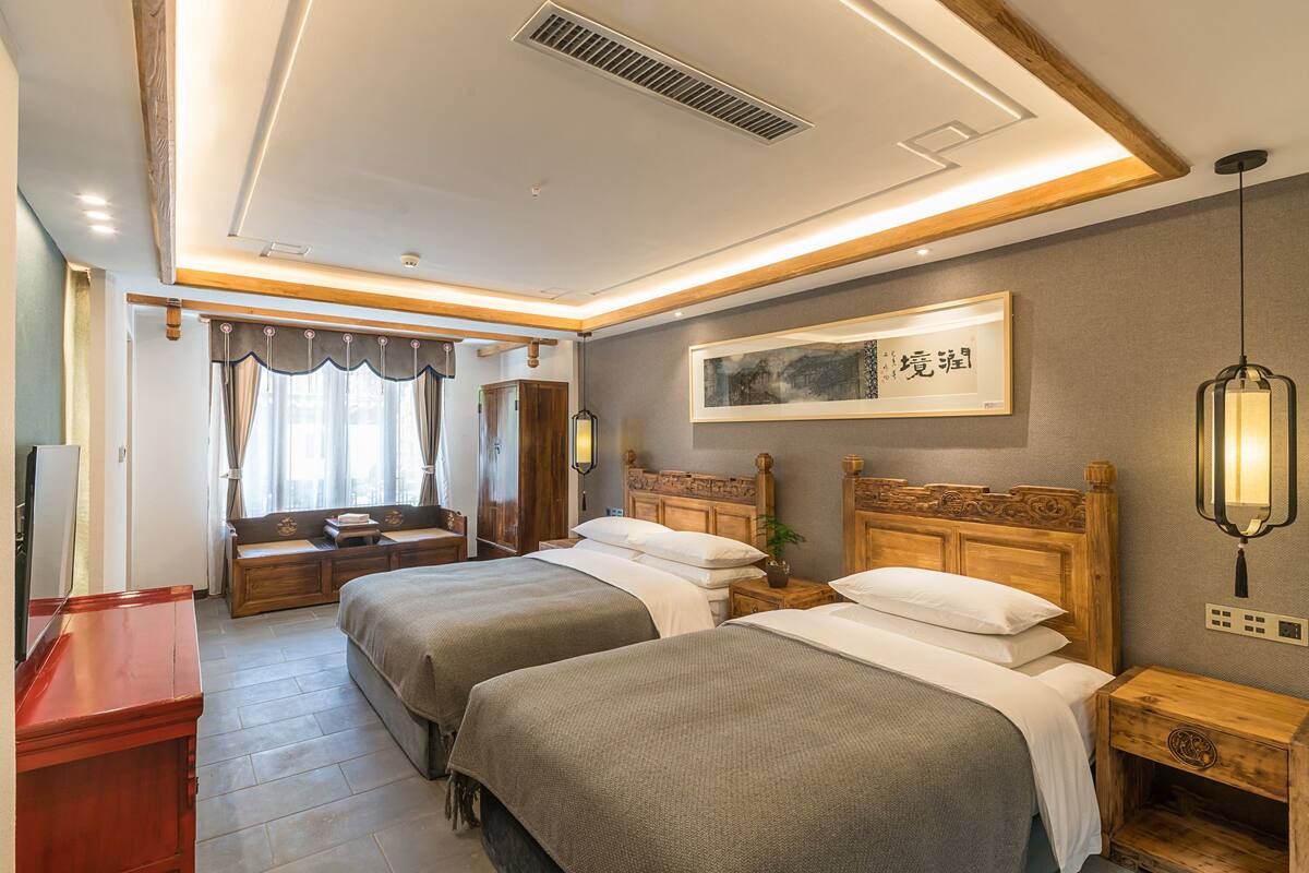 Une chambre d’hôtel avec deux lits, une table de chevet en bois, un bureau et une grande fenêtre avec des rideaux.